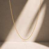 A delicate gold chain necklace draped in a U-shape against a warm beige background with soft diagonal shadows.