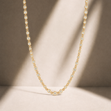 A delicate gold tennis-style chain necklace draped in a U-shape against a warm beige background with soft diagonal shadows.