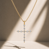 A gold cross pendant necklace adorned with diamond marquise-cut gemstones, suspended from a delicate gold chain against a warm beige background with soft diagonal shadows.