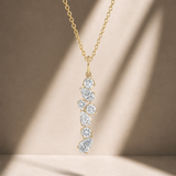 A gold chain necklace with a vertical cluster pendant of clear teardrop-shaped gemstones, displayed against a warm beige background with soft diagonal shadows.