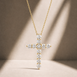 A gold cross pendant necklace adorned with clear gemstones, suspended from a delicate gold chain against a warm beige background with soft diagonal shadows.
