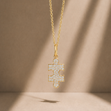 A gold puzzle-piece pendant necklace adorned with small clear pavé stones, suspended from a delicate gold chain against a warm beige background with soft diagonal shadows.